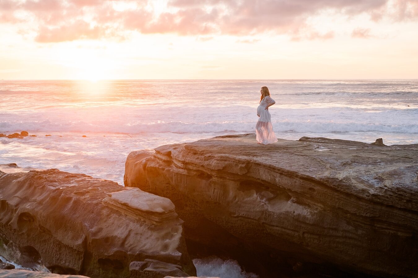 pregnant woman in white dress on beach cliff at sunset | San Diego maternity photographer